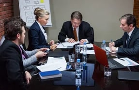 Fotografía de cuatro profesionales de negocios en trajes formales sentados alrededor de una mesa de conferencias con un diagrama de flujo al fondo, analizando métricas de madurez de procesos